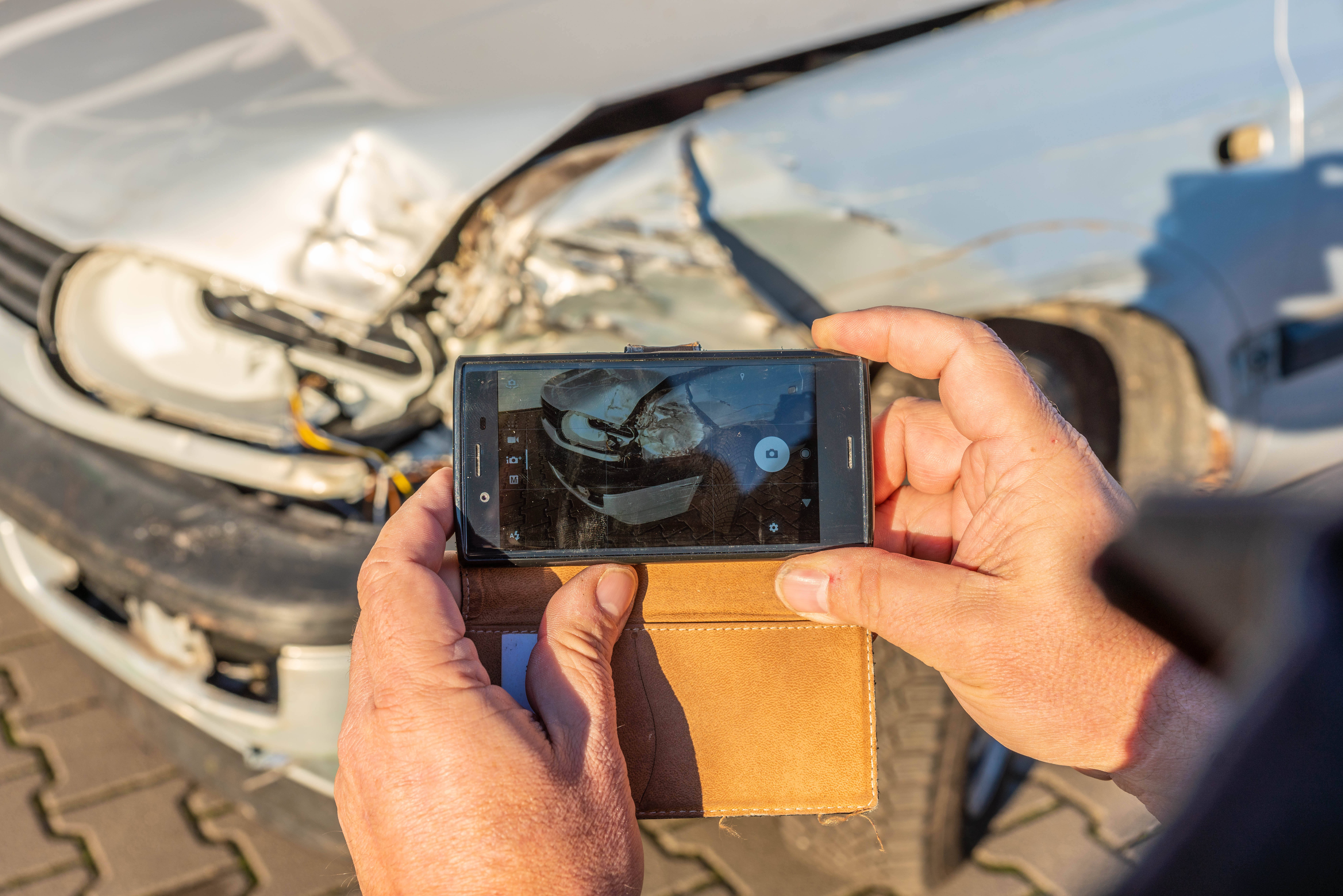 Ein Mann fotografiert mit seinem Smartphone einen Unfallschaden an der Front eines silbernen Autos. Solche Bilder dienen oft als Grundlage für neutrale Kfz-Gutachten, etwa im Rahmen technischer Kfz-Gerichtsgutachten in Hannover.